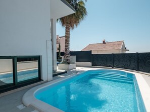 Ein Swimmingpool mit klarem, blauem Wasser, umgeben von einer weißen Poolterrasse, einer Palme und einem Gebäude mit einem Ziegeldach im Hintergrund.
