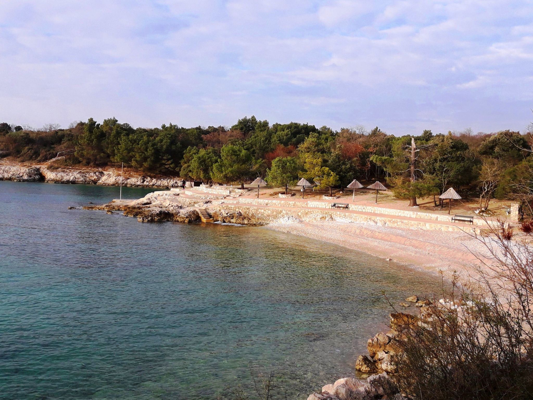 Et kystområde med klart vand, sten trapper ned til en strand og flere strandparasoller.