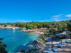 Eine Küstenlandschaft mit klarem, blauem Wasser, einer felsigen Uferlinie sowie mehreren Strandstühlen und Sonnenschirmen.