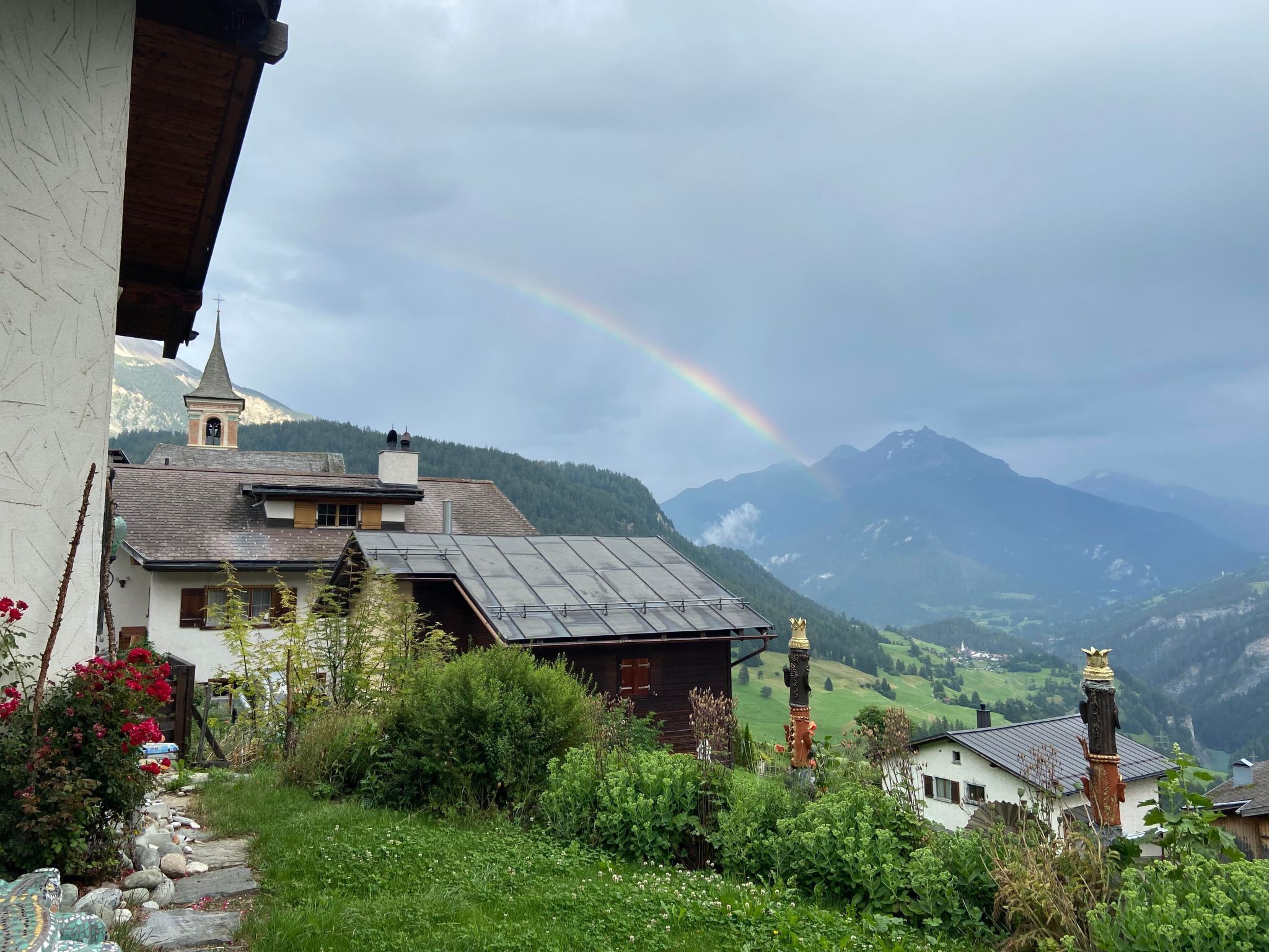 Un village de montagne à l’architecture traditionnelle, une église dotée d’un clocher et un arc-en-ciel surplombant les montagnes.