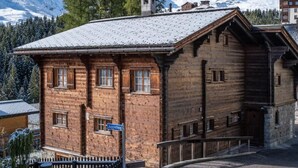 Un edificio tradicional de madera con techo inclinado, situado en una zona montañosa con picos nevados al fondo.