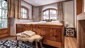 Un dormitorio con una cama de madera, dos ventanas con cortinas, un taburete con una manta de piel y una alfombra estampada.