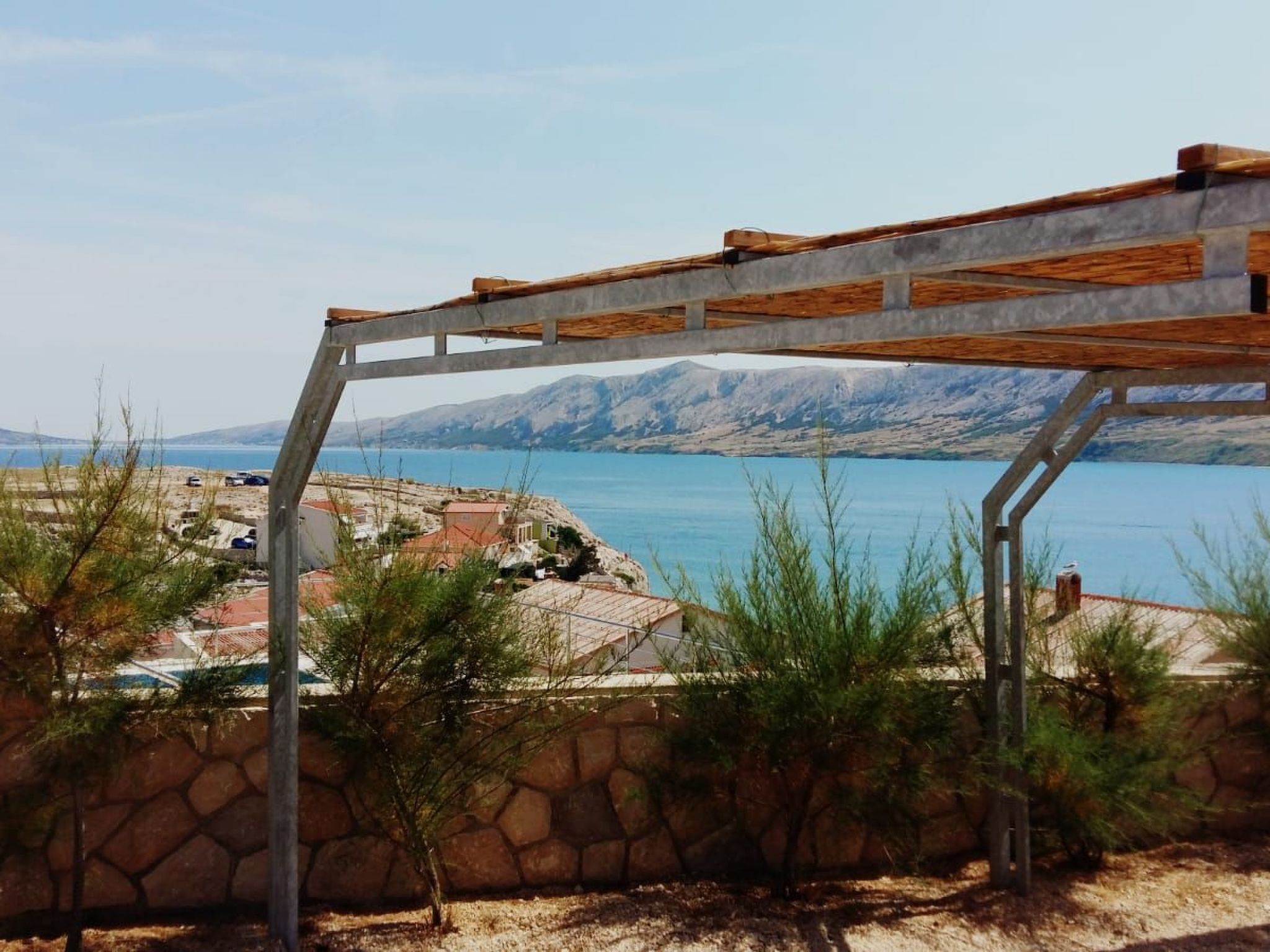Een stenen muur met een pergola die uitkijkt over een kustgebied met huizen en een waterpartij.