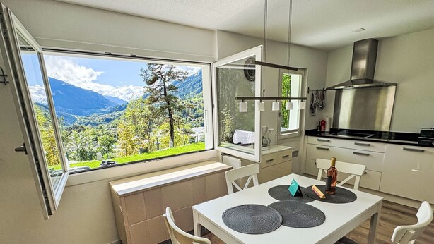 A modern kitchen with a large window, a dining table with chairs, and a view of a mountainous landscape.