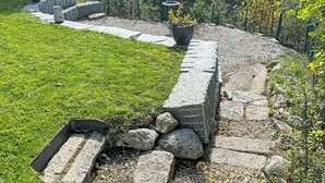 A stone retaining wall with a grassy area and a pathway leading to a garden.