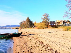 Ein Strand mit einem Ruderboot, sandigem Ufer, Bäumen und Gebäuden im Hintergrund.