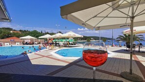 Blick auf ein Schwimmbecken mit einem Glas rotem Getränk, Sonnenschirmen und Personen am Pool.