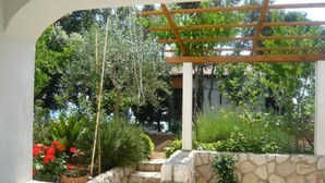 Ein aus Stein gemauerter Garten mit einer Holz-Pergola, Blumentöpfen und verschiedenen Bäumen und Sträuchern.
