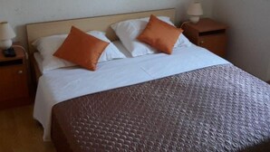 Una cama bien hecha con sábanas blancas y dos almohadas naranjas, un cabecero de madera y una mesita de noche con una lámpara.