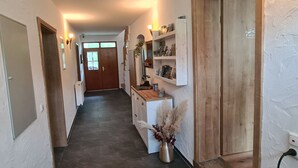 Un pasillo con una puerta de madera al final, una pequeña mesa con un jarrón de plantas secas y una estantería con libros y objetos decorativos.