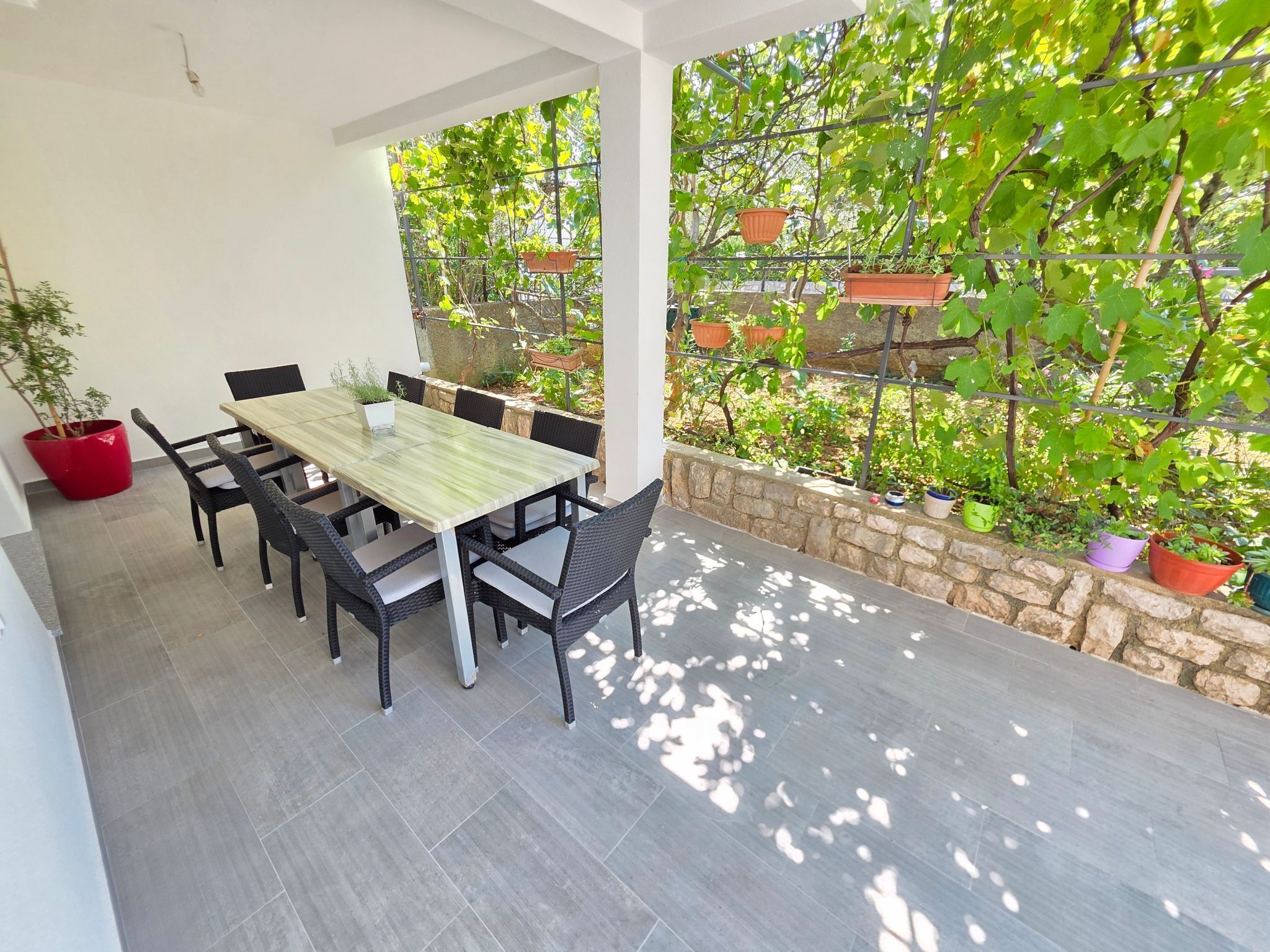En overdækket terrasse med spisebord og stole, omgivet af krukker med planter og en stenvæg.