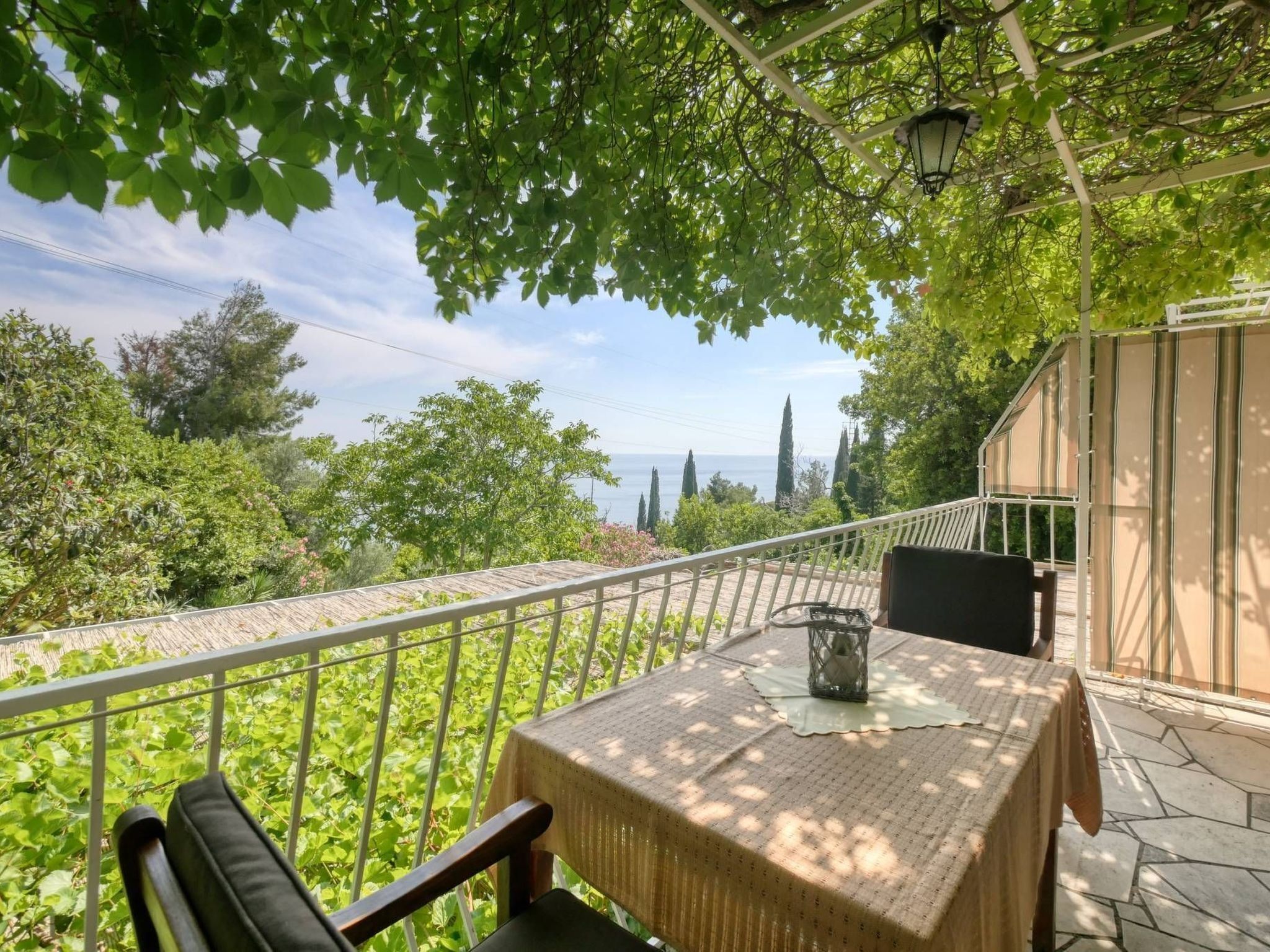 A balcony with a table and chairs, overlooking a lush garden and a distant view of the sea.