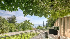 Un balcone con tavolo e sedie, affacciato su un rigoglioso giardino e una vista lontana del mare.