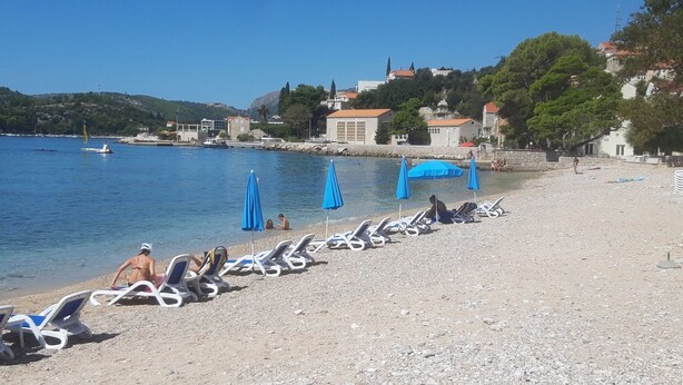 Una spiaggia con ciottoli, lettini e ombrelloni, un mare cristallino e edifici sullo sfondo.