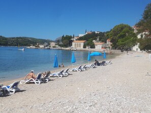 A pebbled beach with sun loungers and umbrellas, a clear blue sea, and buildings in the background. - "Helena2" mit Blick auf das Wasser by Interhome (Dubrovnik/Mlini)