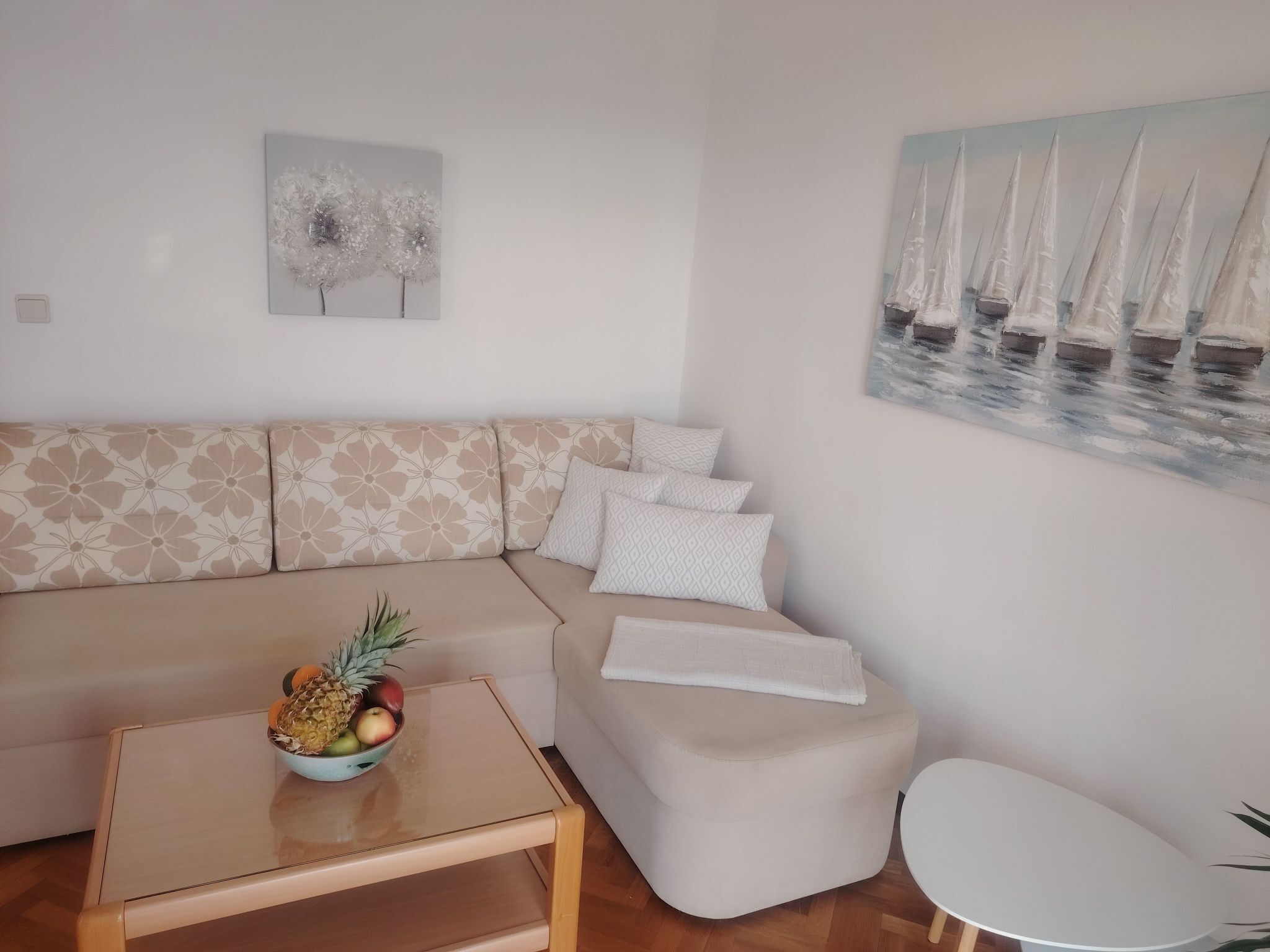 A modern living room with a beige sofa, a glass coffee table with fruit, and a painting of sailboats.