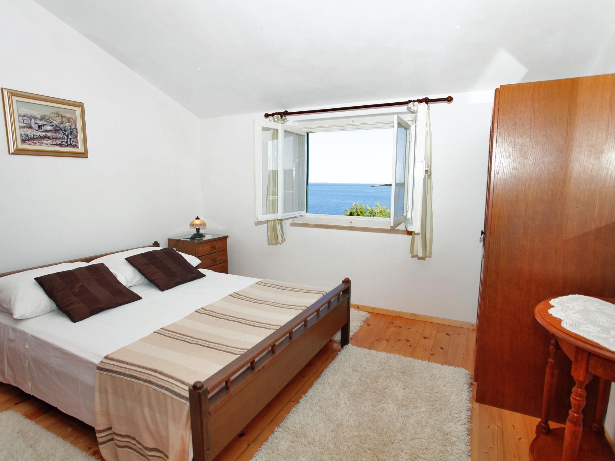 A bedroom with a bed, a nightstand, a window with a view of the sea, and a wooden wardrobe.