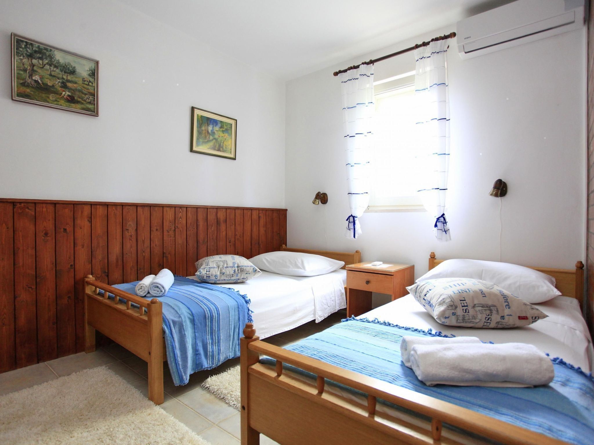 Una habitación con dos camas de madera, ropa de cama blanca y una manta azul. Hay dos cuadros en la pared, una ventana con cortinas y un aire acondicionado instalado en la pared.