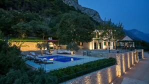 A pool area with lounge chairs, a stone wall, and a building with a red roof. - Villa Miscic by Interhome (Brela)