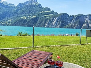 Eine Terrasse mit Blick auf einen See und Berge, ein Holztisch und zwei Gläser roter Flüssigkeit.