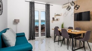 A modern living room with a teal sofa, a dining table with chairs, a TV mounted on the wall, and a view of the sea through the glass door. - Wohnung in Turanj mit Grill by Interhome (Donje Rastane)