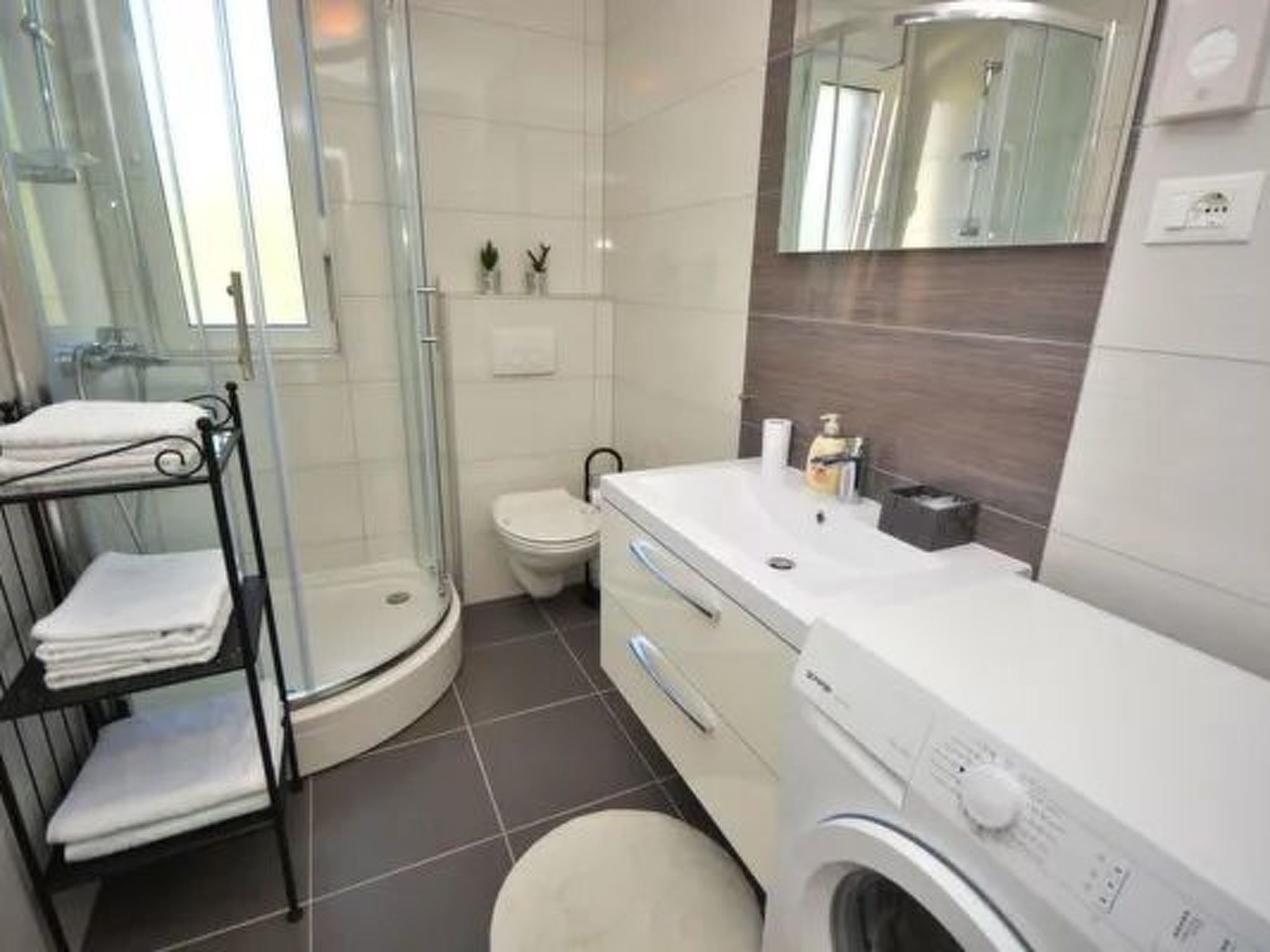 Et moderne badeværelse med brusekabine i glas, toilet, vask med spejl og en indbygget vaskemaskine.