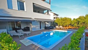 Una casa moderna de dos pisos con piscina rectangular, mobiliario exterior y un jardín bien cuidado.