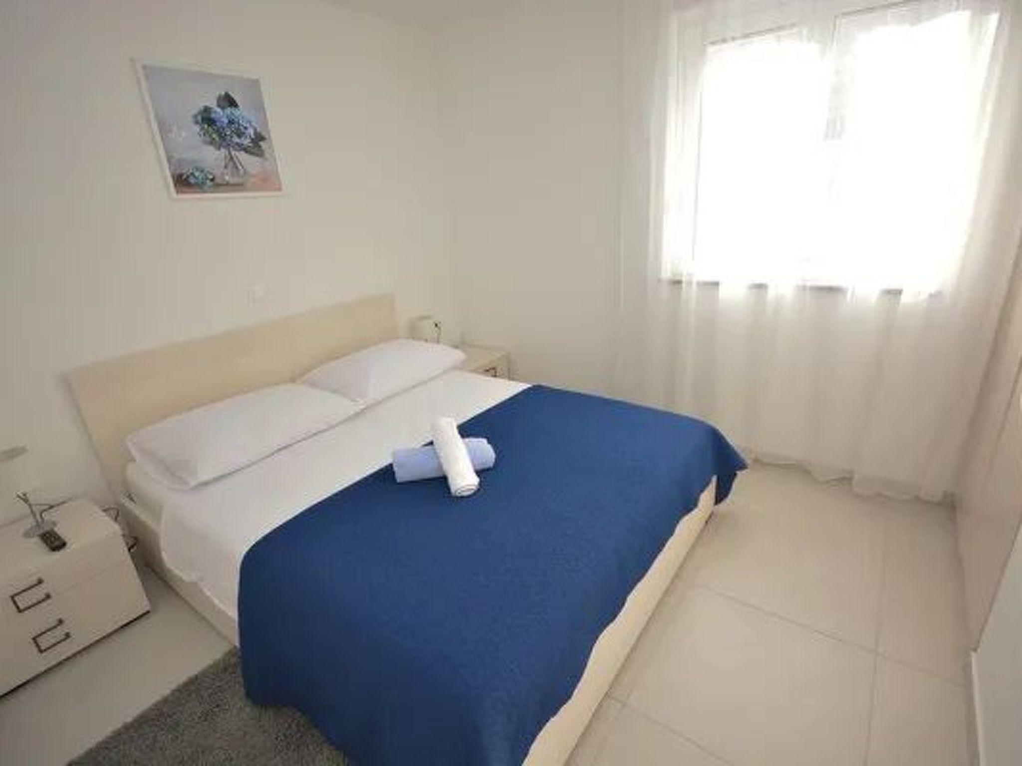 A neatly made bed with a blue blanket, white pillows, and a white headboard. There is a small bedside table with a lamp and a picture frame on the wall above the bed.