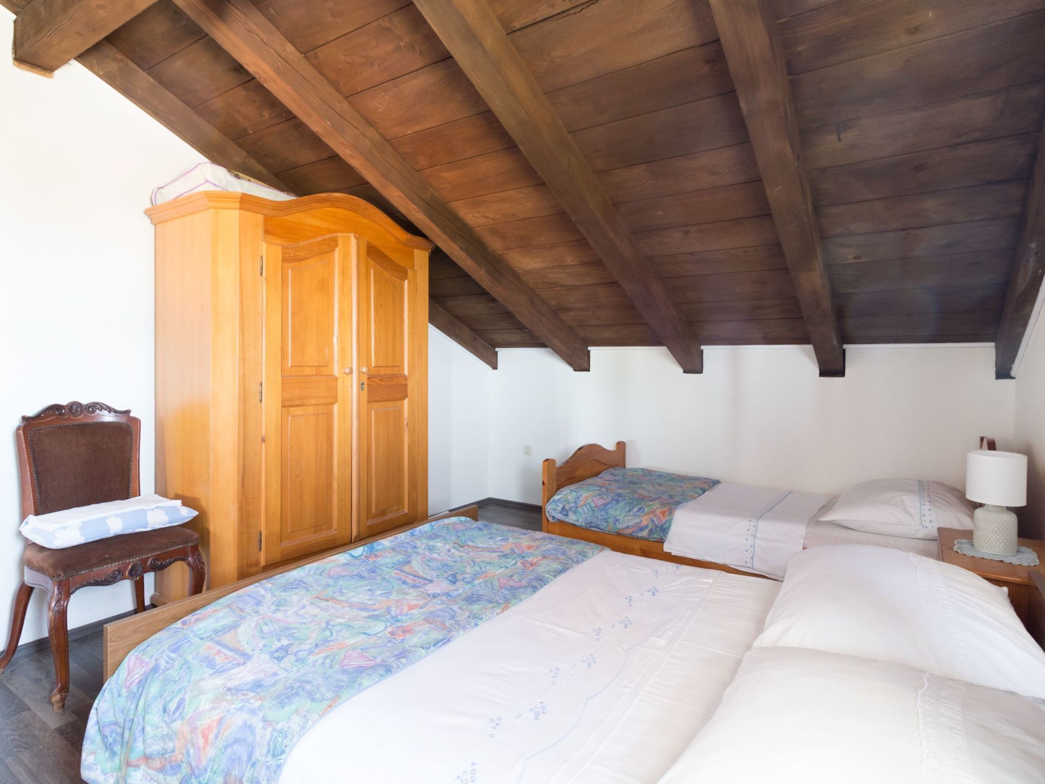 Une chambre avec deux lits, une armoire en bois, une chaise et une table de chevet avec une lampe.