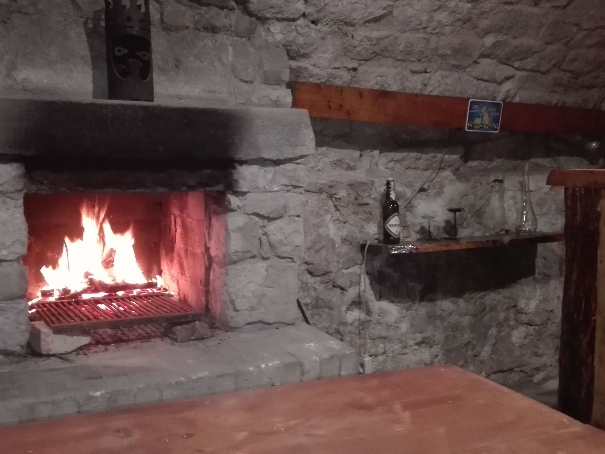 Un camino in pietra con un fuoco acceso, uno scaffale con una bottiglia e dei bicchieri, e una trave di legno sopra.