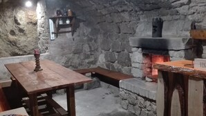 Una habitación rústica con paredes de piedra, una mesa de madera y bancos, una chimenea de piedra y un estante con adornos.