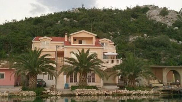 Une grande maison à deux étages, dotée d’un balcon et entourée de palmiers et d’une étendue d’eau.