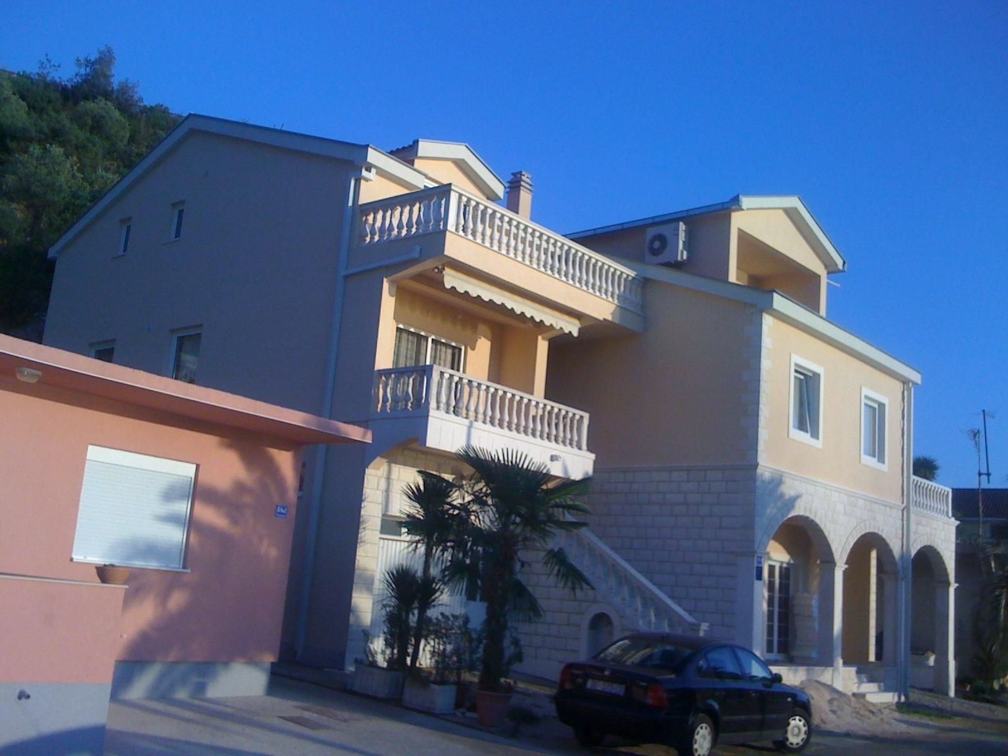 A two-story building with a balcony, a car parked in front, and a palm tree.