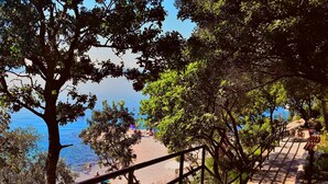 Escadaria de pedra que desce para uma praia, com corrimão, cercada por árvores e com vista para o mar.