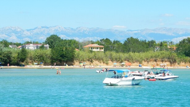 Eine Küstenlandschaft mit Booten auf dem Wasser, einem Strand voller Menschen und Bergen im Hintergrund.