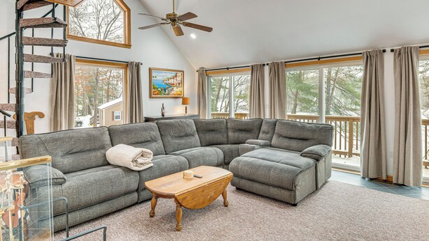 A spacious living room with a large sectional sofa, a wooden coffee table, a spiral staircase, and large windows with curtains. - Waterfront Wakefield Retreat w/ Floating Dock (Sanbornville)