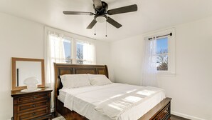 Ein Schlafzimmer mit einem Holzbett, einem Nachttisch, einer Kommode, einem Deckenventilator und zwei mit Vorhängen versehenen Fenstern.