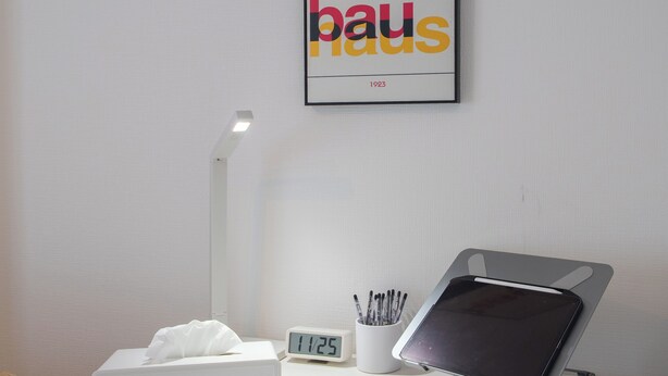 Uma mesa branca com um tablet, uma lâmpada, um relógio e um pôster emoldurado na parede.