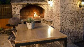 Una chimenea de piedra al aire libre con fuego, una mesa de madera y sillas de mimbre.