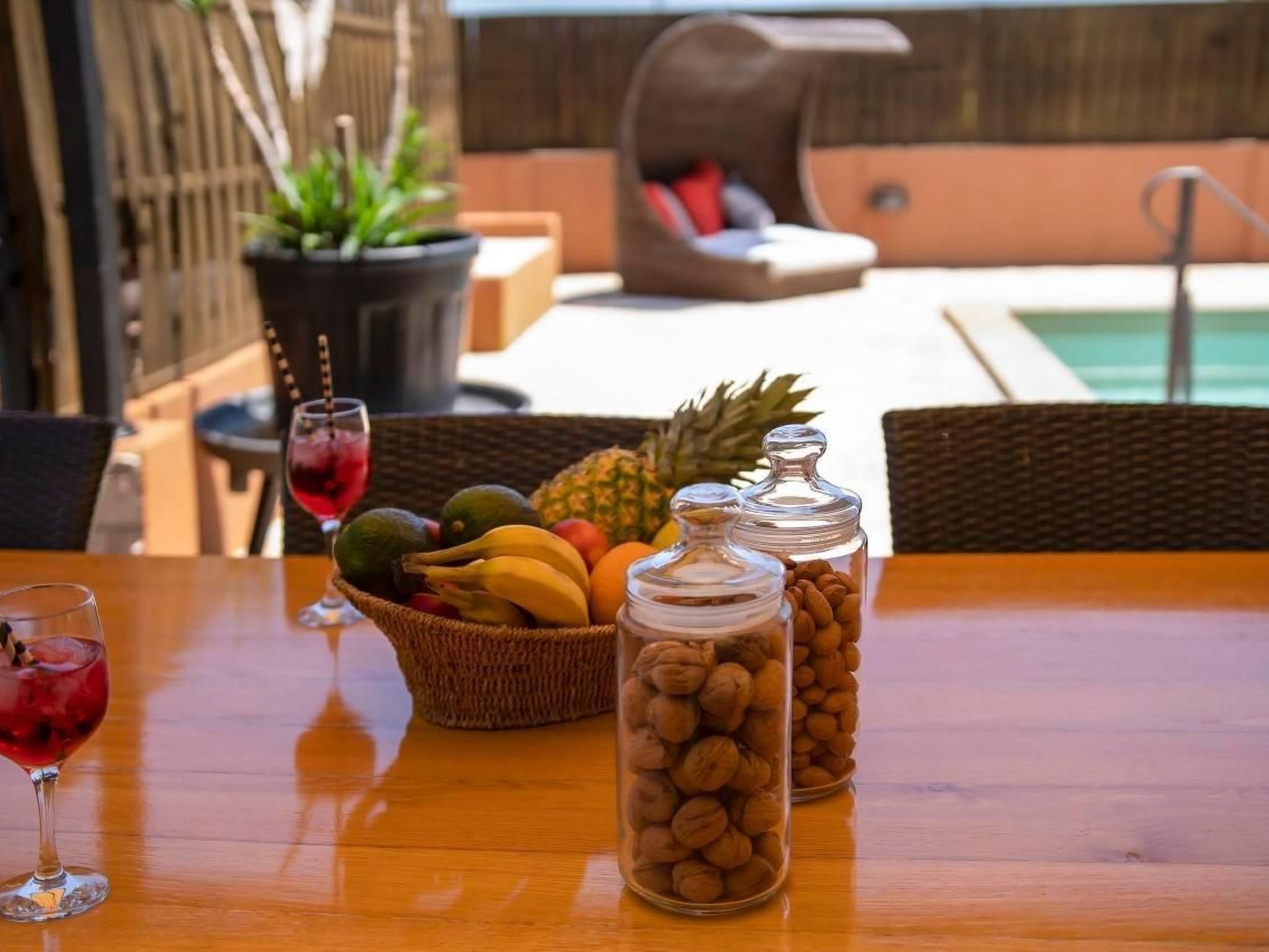 A wooden table with a fruit basket, two glass jars of nuts, and a glass of red drink.