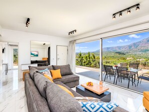 A modern living room with a grey sofa, a coffee table, and a view of a mountainous landscape through large windows. - Luminosa Luxus-Villa by Interhome (Georgioupoli)
