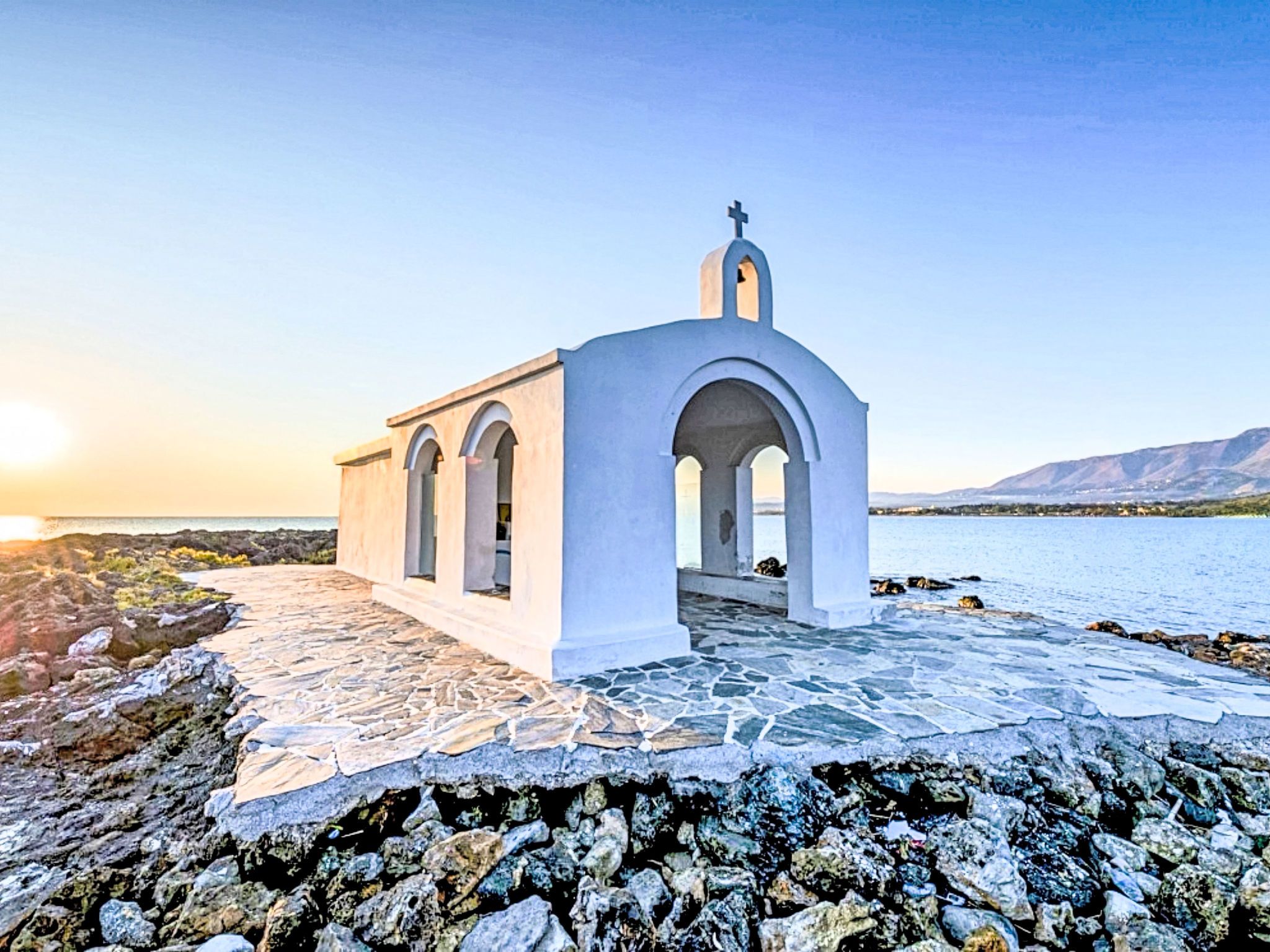 A white church with a cross on top, located near a body of water with mountains in the background.