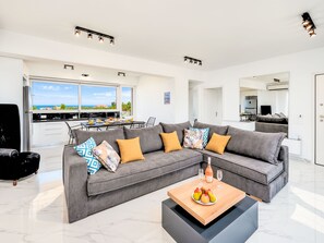 A modern living room with a grey sectional sofa, a coffee table with fruits, and a view of the sea through large windows. - Luminosa Luxus-Villa by Interhome (Georgioupoli)