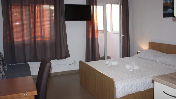 Ein Hotelzimmer mit Bett, Schreibtisch, Sessel, Wandfernseher und einem Fenster mit Vorhängen.