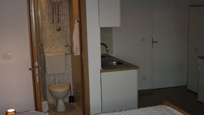 Ein Hotelzimmer mit Bett, Nachttisch mit Lampe, Badezimmer mit Toilette und Waschbecken sowie einer Küchenzeile.