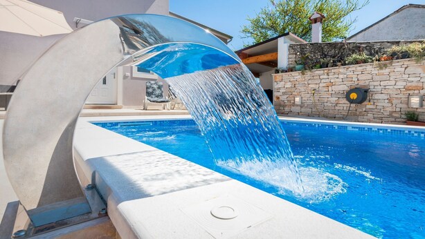 A swimming pool with a water feature, a stone wall, and a house in the background.