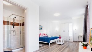 A modern bedroom with a glass-enclosed shower, a bed with a blue bedspread, white wardrobes, and a small white table with a plant.