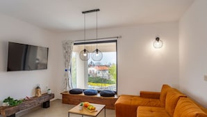 Ein modernes Wohnzimmer mit Sofa, Couchtisch, Fernseher und einem großen Fenster mit Blick auf ein Haus und Bäume.