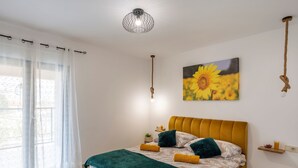 Ein Schlafzimmer mit einem Bett, einem Sonnenblumenbild, einer Pendelleuchte und einem Fenster mit Vorhängen.