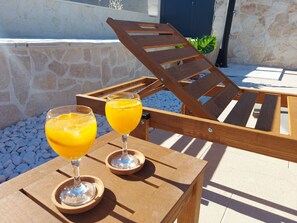 Een houten lounge stoel met een kussen, twee glaasjes jus d'orange op een houten tafel, en een stenen muur op de achtergrond.
